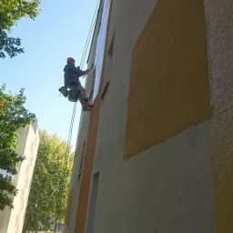 Pracownik na linach maluje elewację bloku z geometrycznym wzorem, widoczny świeżo pomalowany fragment ściany, zabezpieczone okna, drzewa w tle.