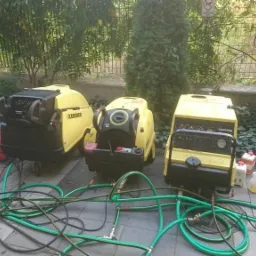 Trzy żółte myjki ciśnieniowe Kärcher z podłączonymi zielonymi wężami stoją na chodniku, w tle zieleń krzewów i drzew.