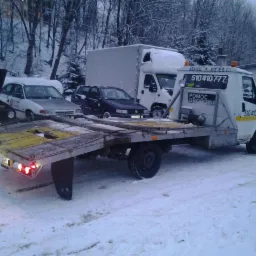 Laweta z napisem 'Pomoc Drogowa' na śniegu, z załadowanymi autami. Widoczne opony w tle i drzewa pokryte śniegiem. Zima. Numer telefonu na kabinie.