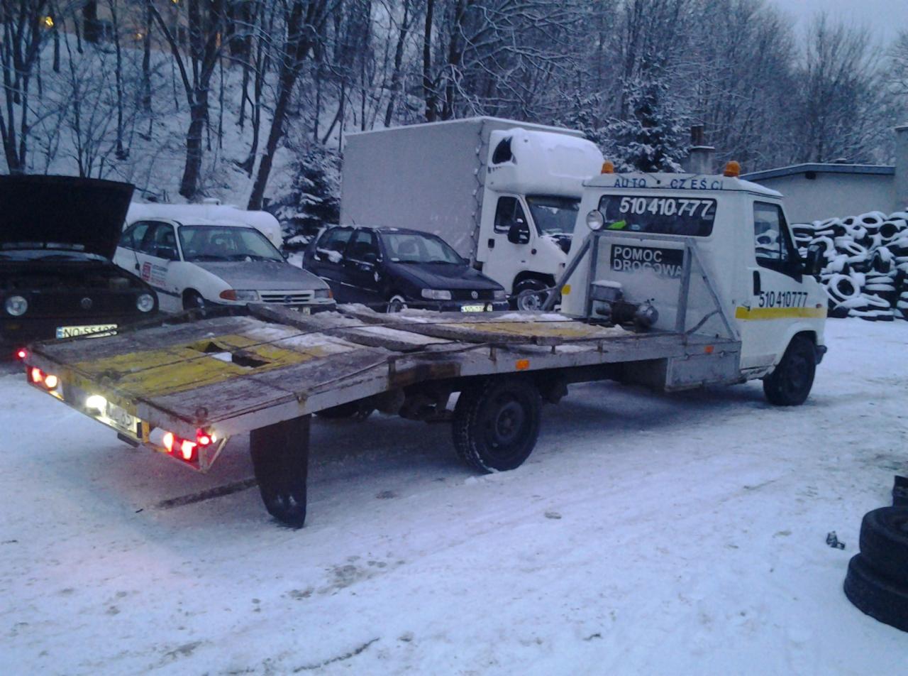 Laweta z napisem 'Pomoc Drogowa' na śniegu, z załadowanymi autami. Widoczne opony w tle i drzewa pokryte śniegiem. Zima. Numer telefonu na kabinie.