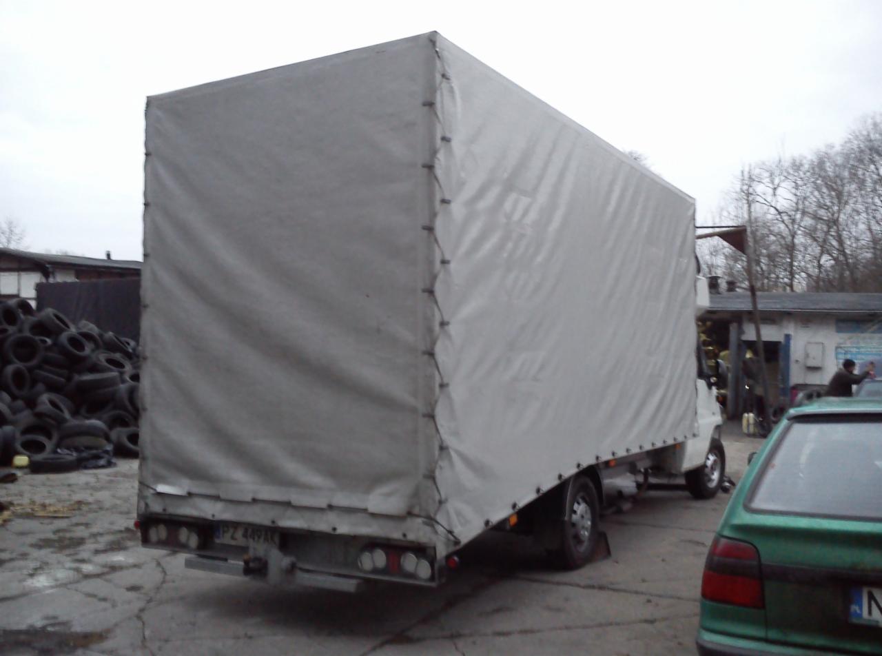 Biały samochód dostawczy z plandeką zaparkowany na placu, widok z tyłu. W tle sterta opon i budynek warsztatowy.