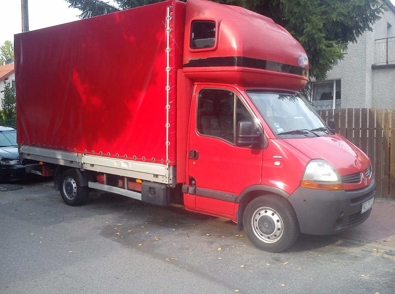 Czerwony samochód dostawczy Renault Master z plandeką na pace, zaparkowany na ulicy w słoneczny dzień. Widoczny fragment budynku w tle.