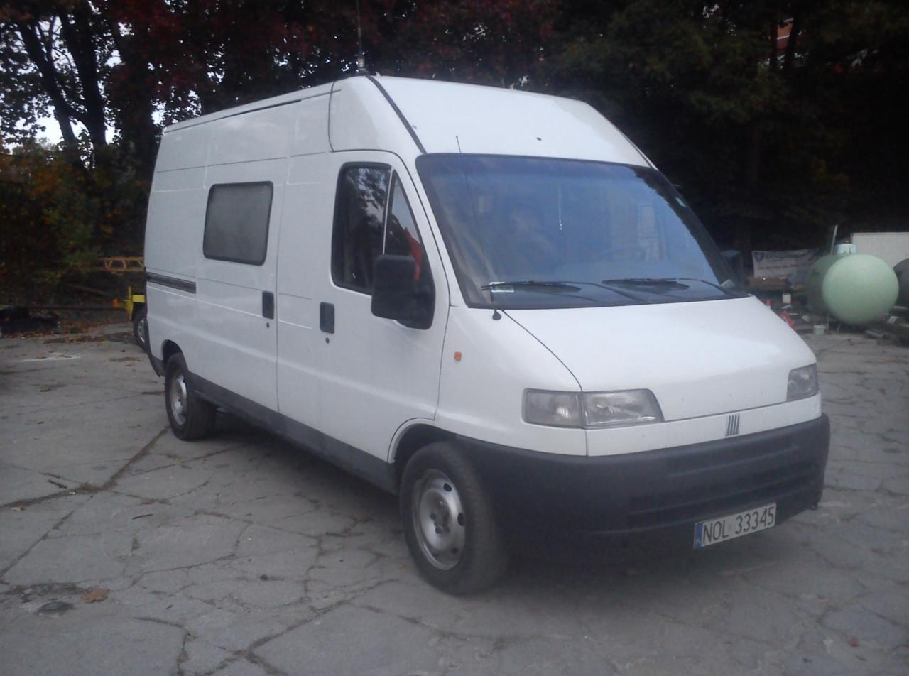 Biały bus dostawczy z anteną na dachu, zaparkowany na betonowym placu, widok z boku na tle zieleni. Tablica rejestracyjna widoczna.