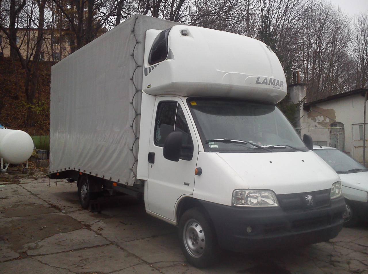 Biały samochód dostawczy marki Peugeot z plandeką i napisem LAMAR, zaparkowany na zewnątrz w pochmurny dzień. Widoczny zbiornik i budynek w tle.