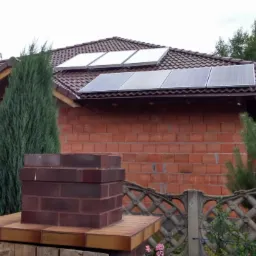 Panele fotowoltaiczne na dachu domu z czerwonej cegły, widoczne z poziomu ogrodu z tujami i betonowym płotem. Częściowo widoczny komin z cegły.