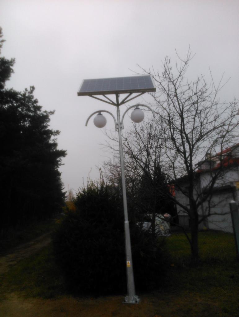 Lampa solarna parkowa z dwoma kulistymi kloszami, zamontowana na zewnątrz, w tle drzewa i budynek. Szary, pochmurny dzień.