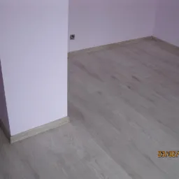Pomalowane na liliowo ściany i ułożona jasna podłoga laminowana z drewnopodobnym wzorem, widoczne listwy przypodłogowe i gniazdko elektryczne.
