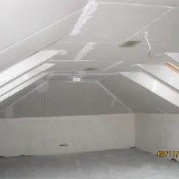 Poddasze z zamontowanymi płytami gipsowo-kartonowymi, widoczne szpachlowane łączenia, instalacja elektryczna i okno dachowe w trakcie wykańczania.