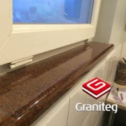 Czerwono-brązowy, polerowany parapet granitowy pod białym oknem PCV, widoczne logo 'Graniteg' w prawym dolnym rogu.