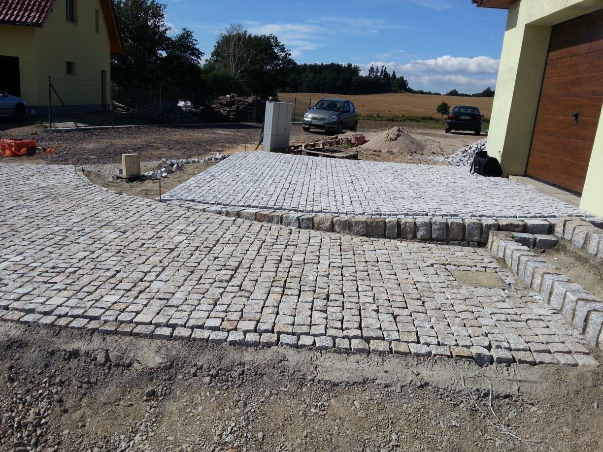 Układanie kostki granitowej w Lubiechowie na posesji prywatnej, widoczne fragmenty ułożonej nawierzchni i przygotowane podłoże, w tle budynek mieszkalny i samochody.