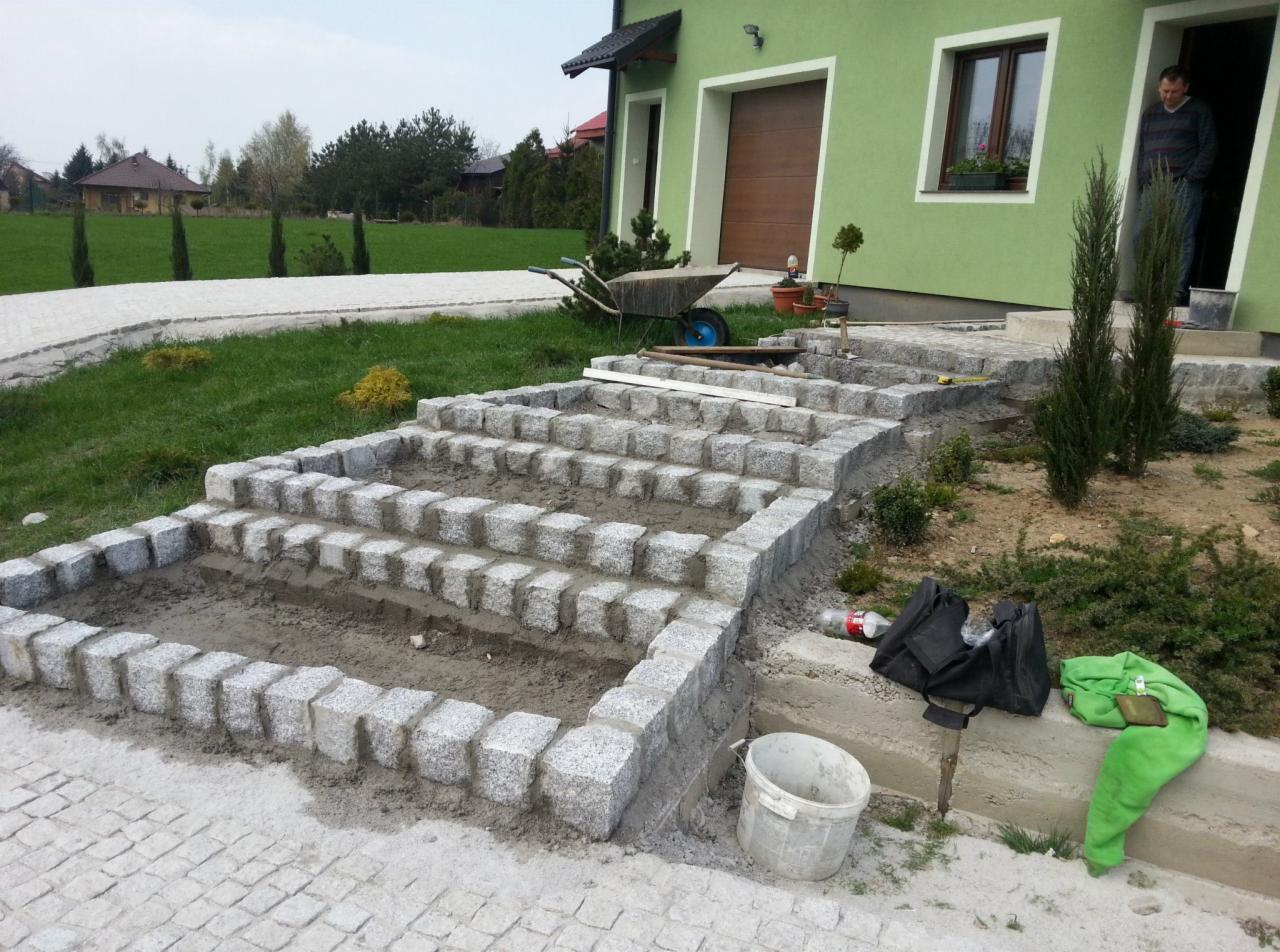 Budowa schodów zewnętrznych z kostki granitowej na posesji w Słotwinie, widoczna warstwa podsypki i częściowo ułożona kostka, w tle zielony dom z drewnianymi drzwiami garażowymi i pracownik w progu.