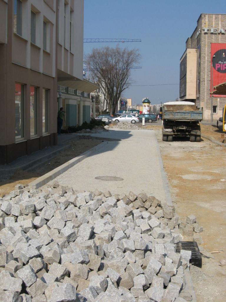 Stos szarych kostek granitowych na placu budowy w Radomiu, w tle budynek Opery i ciężarówka z piaskiem.