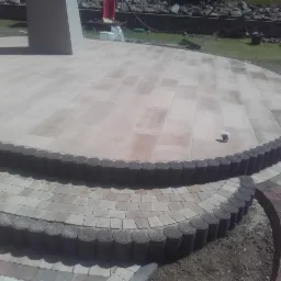 Taras z kostki brukowej o zaokrąglonym kształcie, z dwoma stopniami obłożonymi kostką i obrzeżem z ciemnych, okrągłych elementów. Widoczny fragment ściany budynku oraz trawnik w tle.