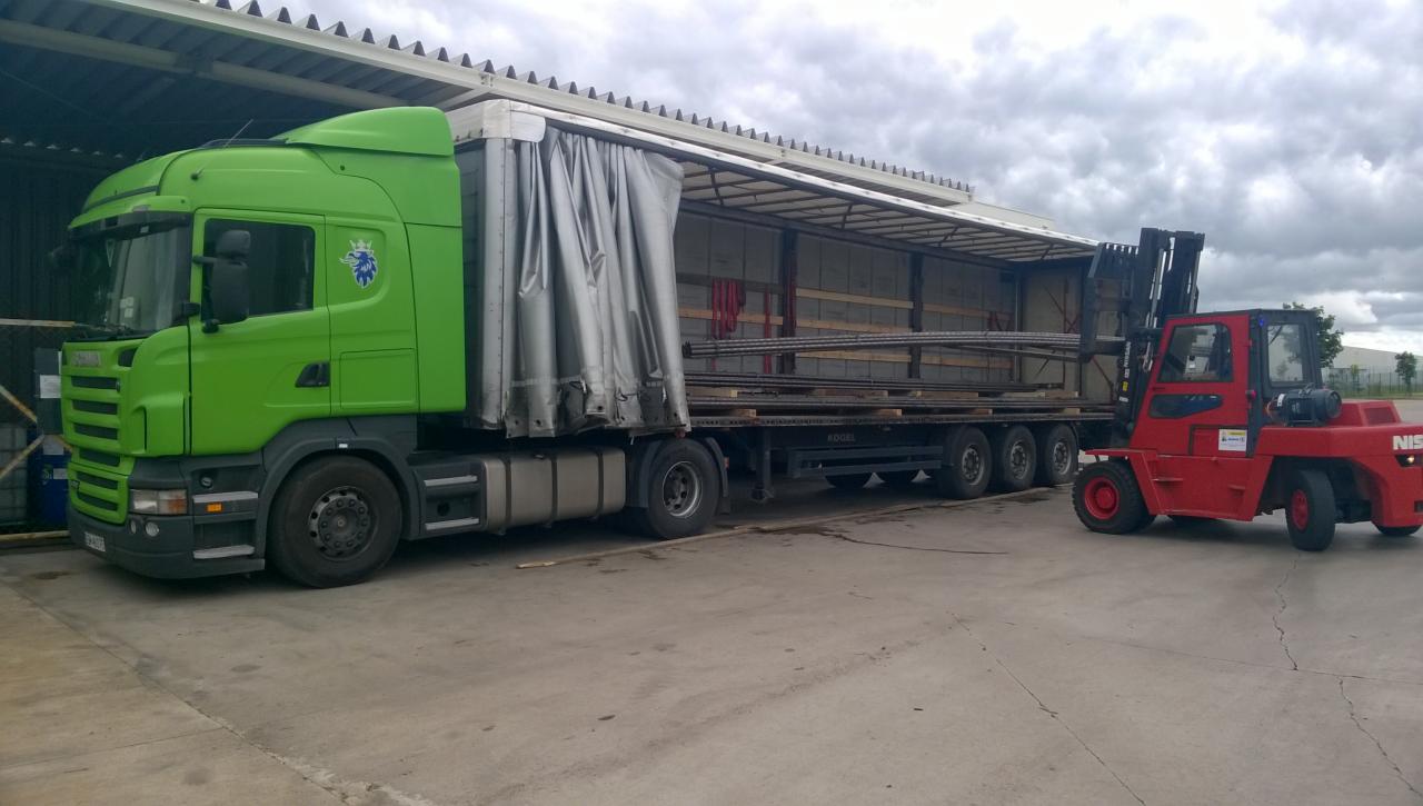 Zielona ciężarówka Scania z naczepą częściowo odsłoniętą, załadowana metalowymi prętami, obok czerwony wózek widłowy Nissan gotowy do rozładunku, pod zadaszeniem magazynu.