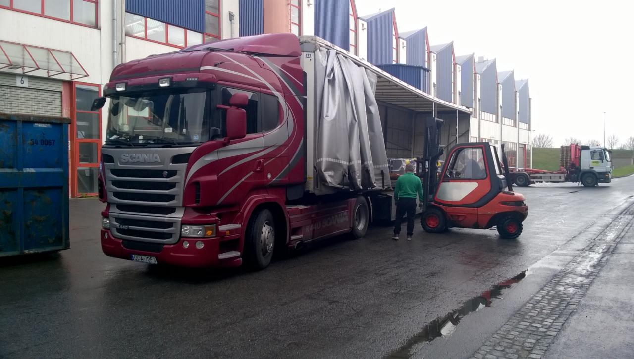 Czerwona ciężarówka Scania z naczepą częściowo odsłoniętą, obok wózek widłowy i pracownik w zielonej bluzie, na mokrym placu przed halą magazynową.