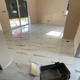 Wykończone płytki podłogowe z marmurowym wzorem w jasnym, przestronnym pomieszczeniu. Widoczny wałek malarski i kuweta malarska. Ściany pomalowane na jasny, beżowy kolor. Duże okno z roletami.