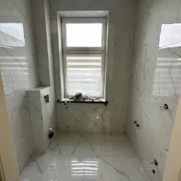 Wnętrze łazienki w trakcie prac wykończeniowych, ściany i podłoga wyłożone białymi płytkami imitującymi marmur, widoczne przygotowane punkty instalacyjne, okno z żaluzjami i narzędzia na parapecie.