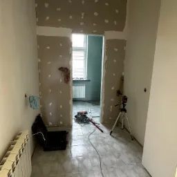 Wnętrze w trakcie prac: fragment ściany z płyt kartonowo-gipsowych z widocznymi punktami szpachlowania, poziomica laserowa na statywie, narzędzia i płytki podłogowe.