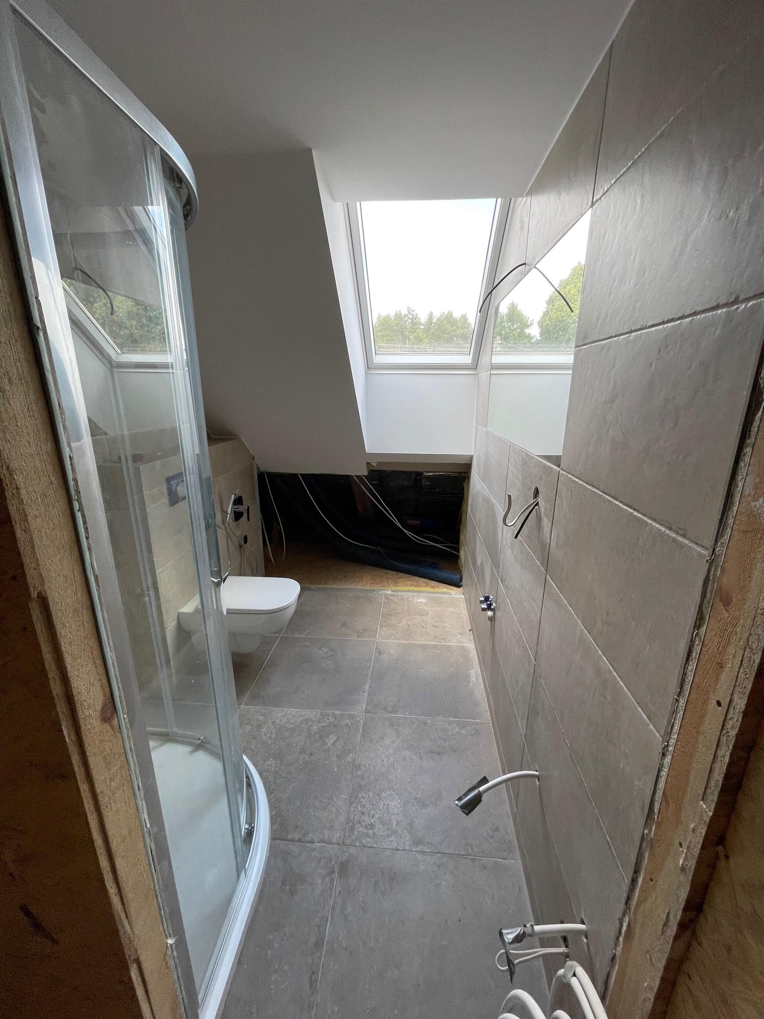 Wnętrze łazienki w trakcie remontu w Międzychodzie: widoczna kabina prysznicowa, toaleta, płytki na ścianach, okno dachowe i odsłonięte przewody.