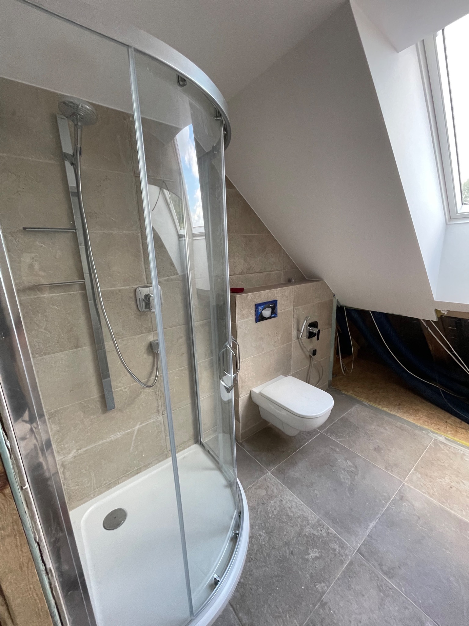 Ukończona łazienka na poddaszu ze skośnym sufitem, z kabiną prysznicową z zaokrąglonymi drzwiami, wiszącą toaletą i płytkami w odcieniach beżu i szarości.