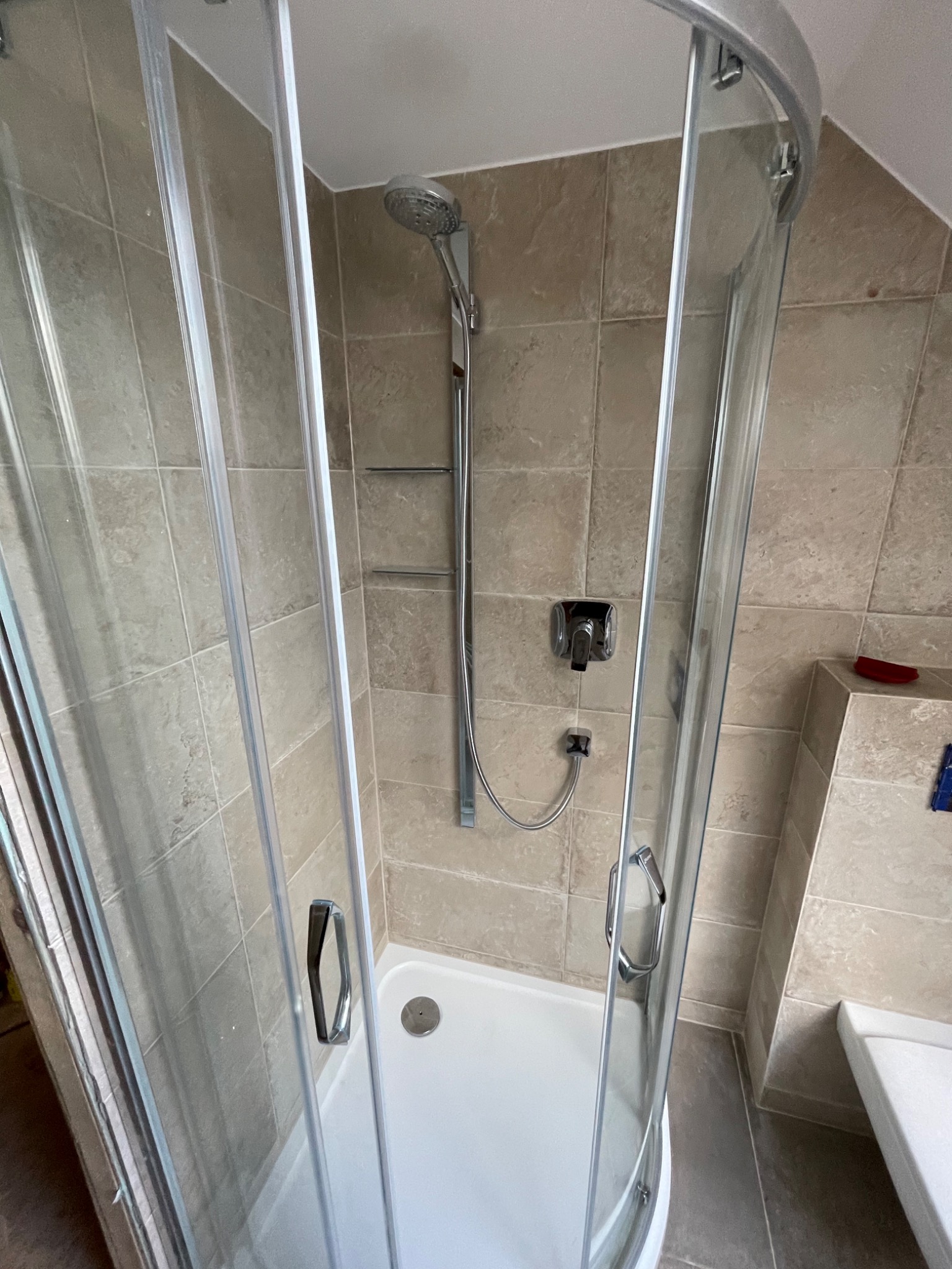 Kabina prysznicowa z jasnymi, kwadratowymi płytkami na ścianach, chromowaną armaturą i białym brodzikiem, widziana z perspektywy lekko z góry.