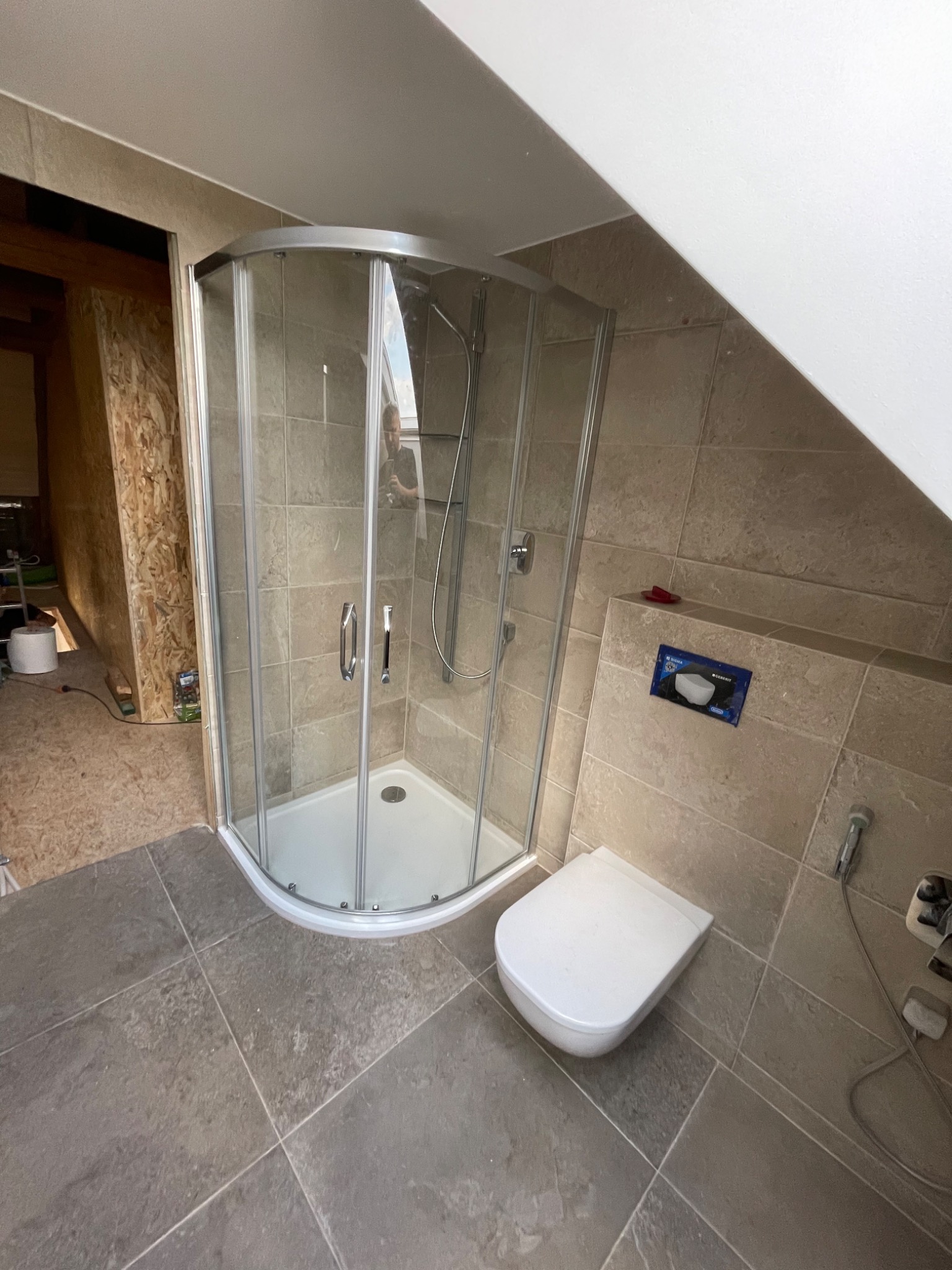 Wykończona łazienka na poddaszu ze skośnym sufitem, z kabiną prysznicową z zaokrąglonym szkłem, podwieszaną toaletą i beżowymi płytkami na ścianach i szarymi na podłodze. W tle widoczna surowa...