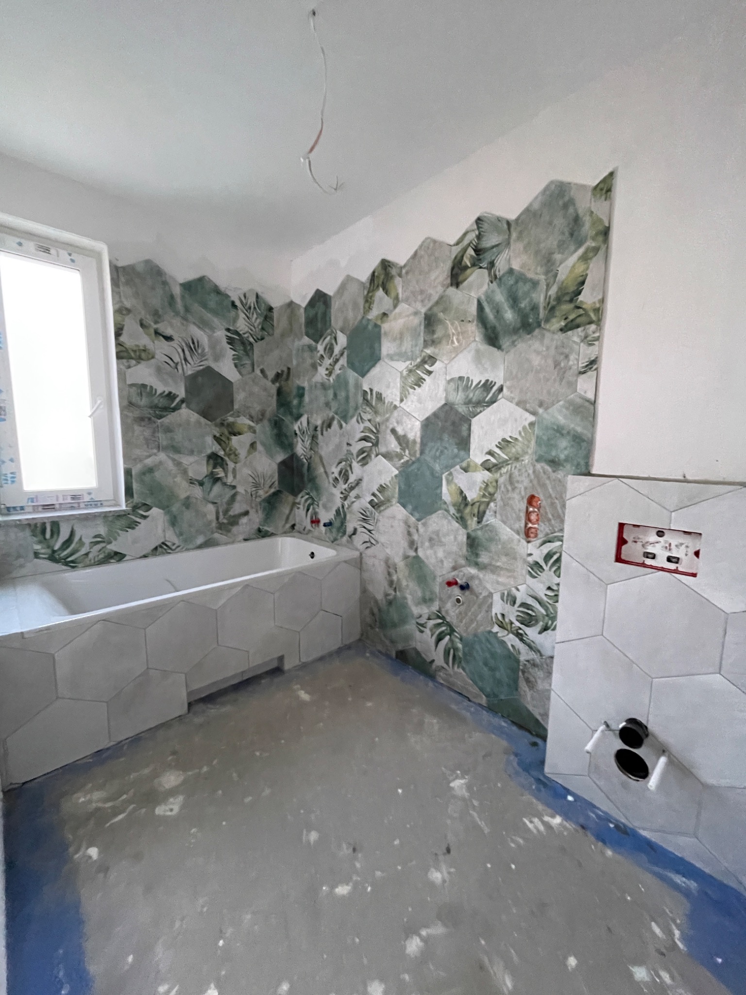 Łazienka w trakcie remontu: heksagonalne płytki z motywem liści na ścianie, obudowa wanny z heksagonalnych płytek, widoczne instalacje hydrauliczne, surowy beton na podłodze.