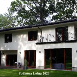 Dwukondygnacyjny, nowoczesny dom z płaskim dachem i balkonem z czarną balustradą, otoczony zielenią drzew i trawnikiem.