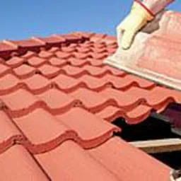 Układanie czerwonej dachówki falistej na dachu, widoczne rękawice robocze i fragment nieba. Dachówka ceramiczna, etap prac dekarskich.