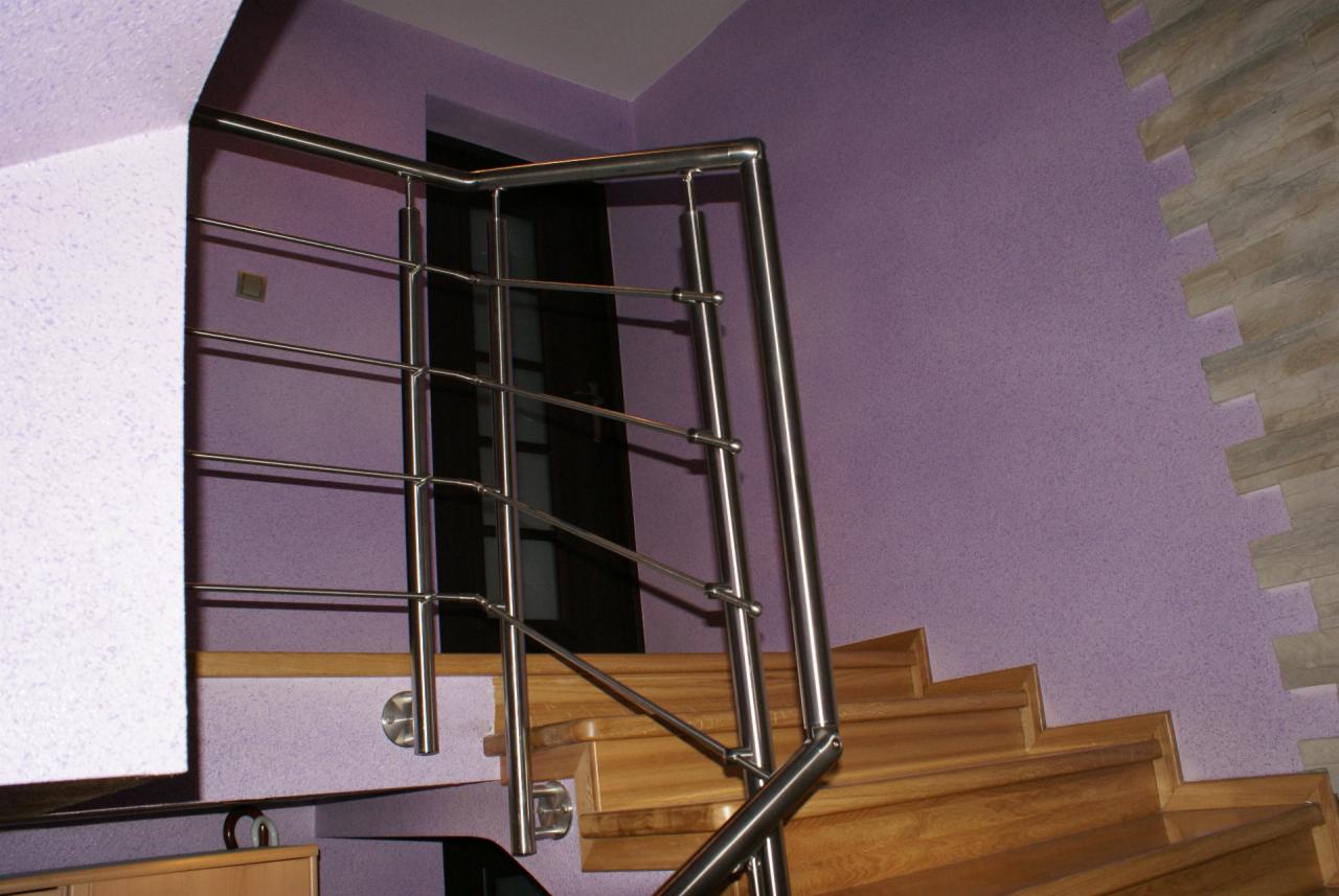 Metalowa balustrada ze stali nierdzewnej, zamontowana na drewnianych schodach. Fioletowa ściana z dekoracyjną okładziną w tle. Detal nowoczesnego wykończenia wnętrza.