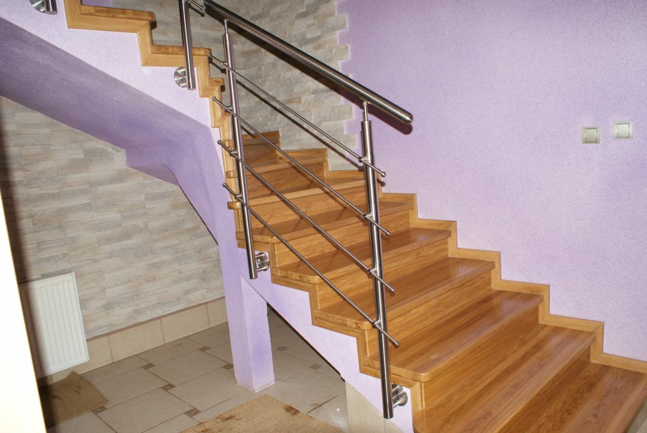 Klatka schodowa z drewnianymi stopniami i metalową balustradą w nowoczesnym domu. Ściany w odcieniach fioletu i beżu. Widoczny grzejnik i fragment podłogi.