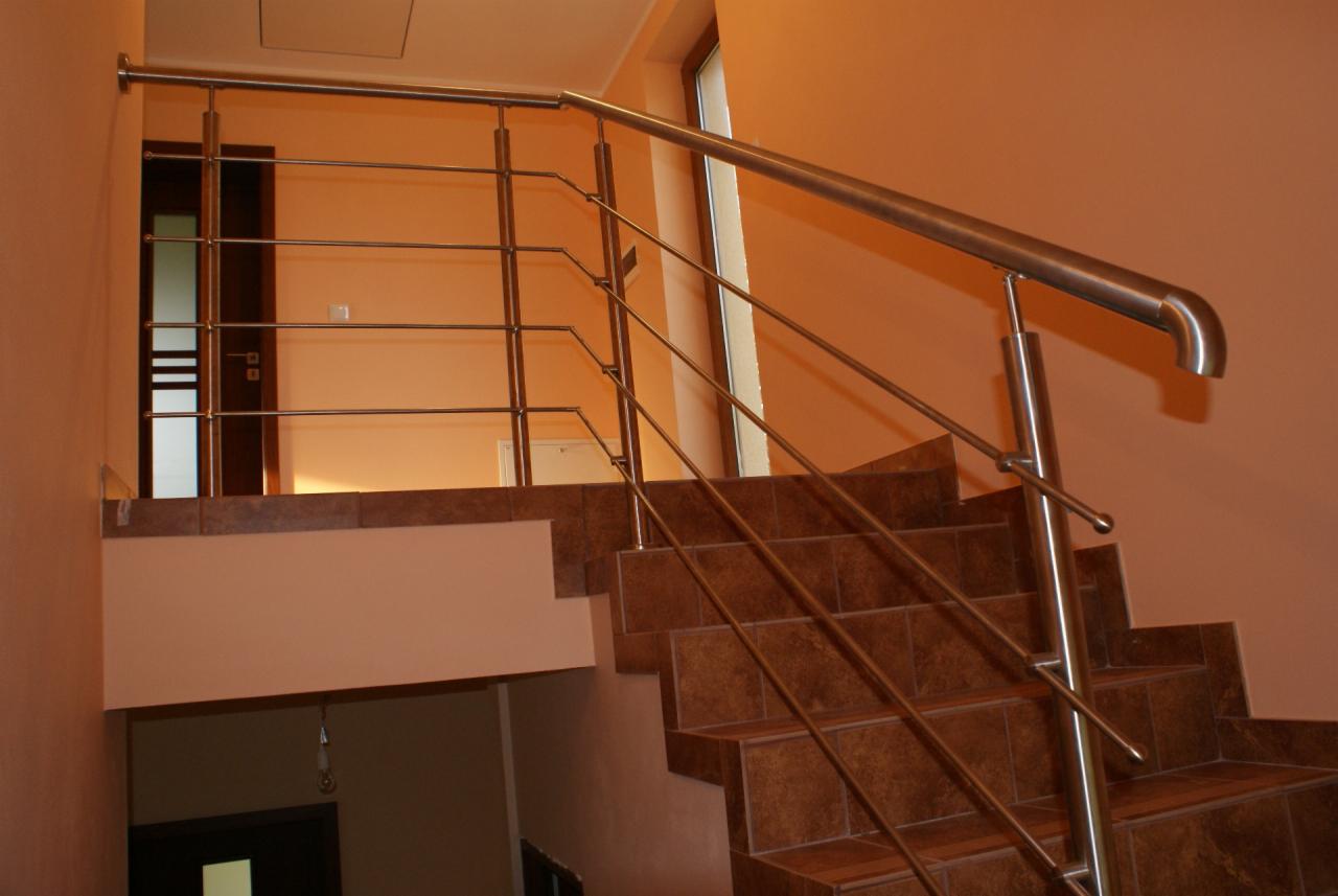 Nowoczesna, metalowa balustrada schodowa ze stali nierdzewnej, zamontowana wewnątrz budynku. Brązowe płytki na schodach, pomarańczowe ściany. Widok z dołu na balustradę.