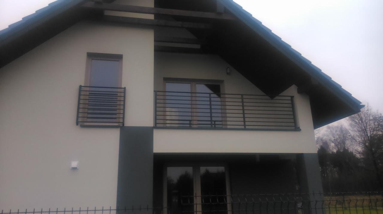 Nowoczesny dom z balkonami zabezpieczonymi metalowymi, poziomymi barierkami. Minimalistyczny design, szara kolorystyka elewacji. Widok z dołu, fragment ogrodzenia.