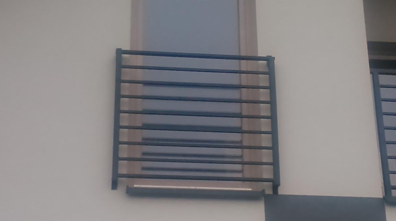 Nowoczesna, pozioma balustrada okienna z metalu, zamontowana na elewacji budynku. Minimalistyczny design, szare elementy, widok z zewnątrz.