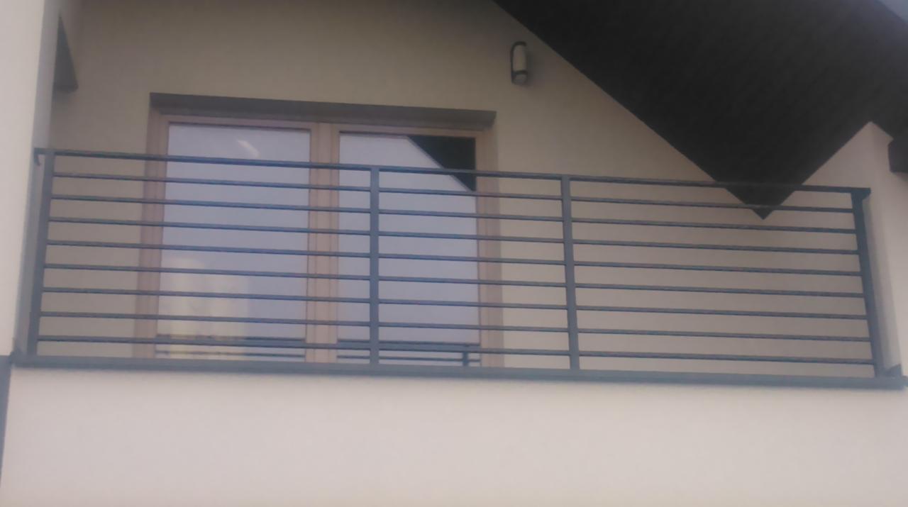 Nowoczesna, metalowa balustrada balkonowa z poziomymi prętami, zamontowana na jasnej elewacji budynku z widocznym oknem balkonowym i fragmentem dachu.