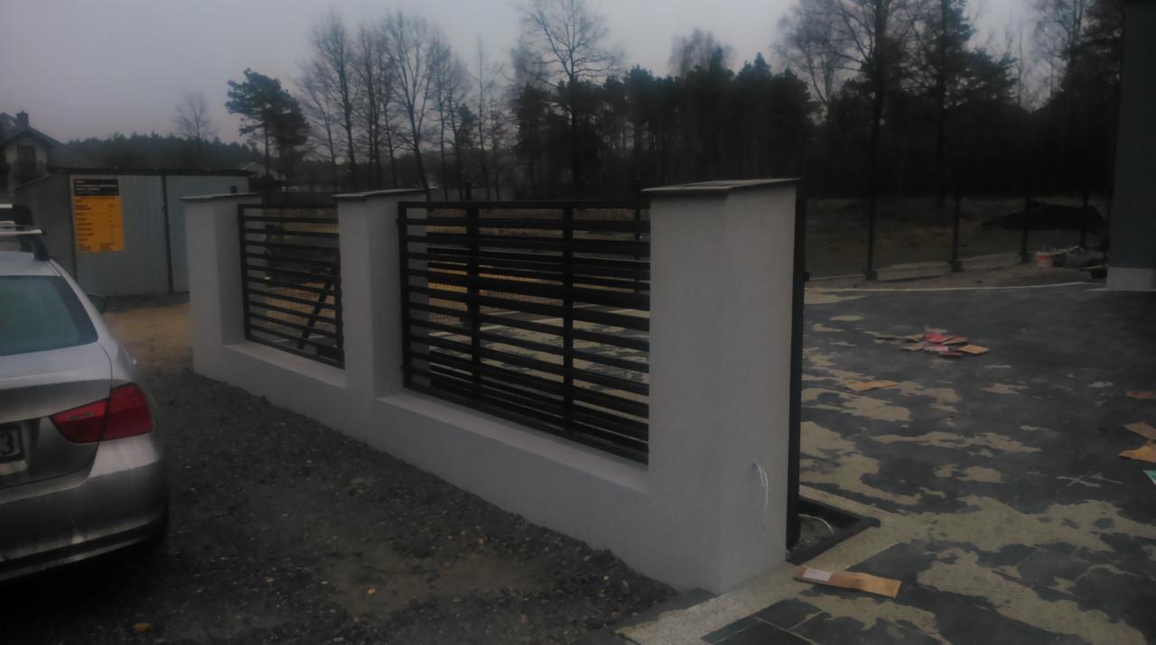 Nowoczesne ogrodzenie z poziomych, czarnych profili, osadzone na niskim, białym murku. Widok z boku, z fragmentem samochodu po lewej stronie. Tło stanowią drzewa.