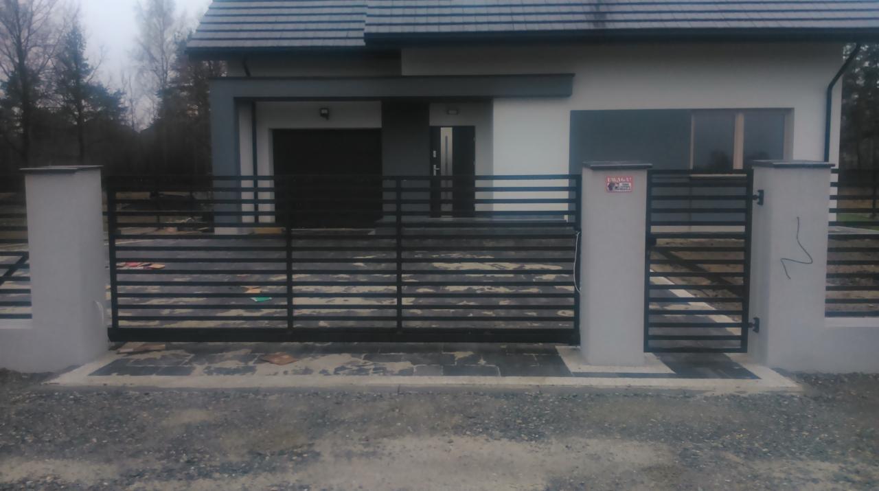 Nowoczesna, pozioma brama przesuwna z czarnych profili, osadzona między szarymi słupkami na tle domu z garażem. Widoczny fragment ogrodzenia.