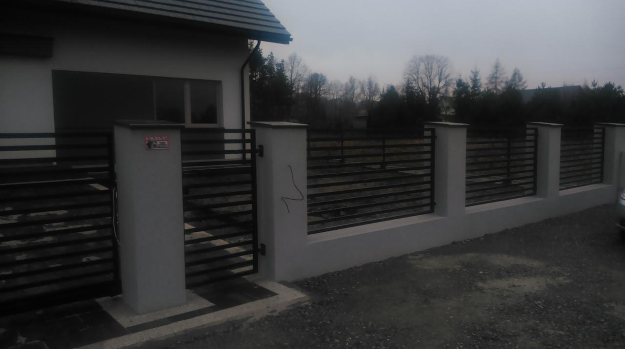 Nowoczesne ogrodzenie posesji z poziomymi, czarnymi przęsłami i białymi słupkami, widoczne z zewnątrz. Na jednym ze słupków drobne graffiti.