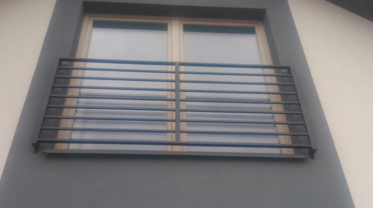 Stylowa, pozioma, metalowa balustrada okienna w kolorze grafitowym, zamontowana na elewacji domu. Widok od dołu, okno z drewnianą ramą w tle.