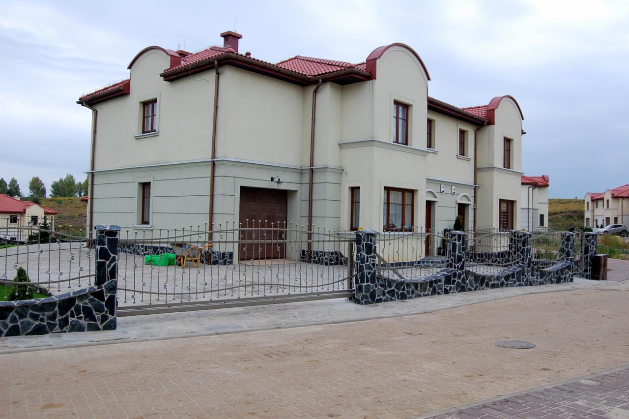 Elegancka, przesuwna brama z metaloplastyki, osadzona na kamiennych słupkach, przed domem z garażem i czerwoną dachówką. Widok z poziomu ulicy.