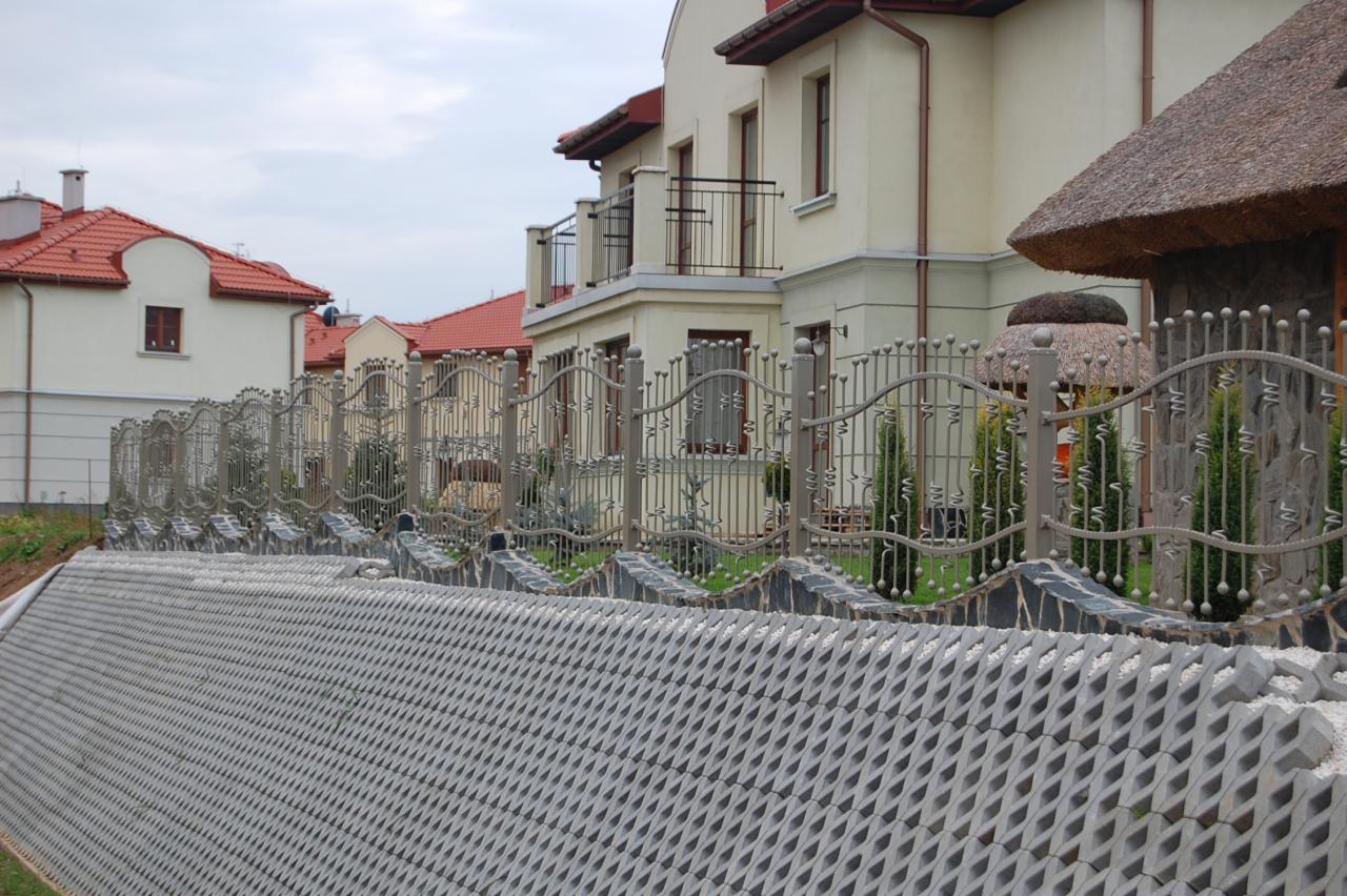 Metalowe ogrodzenie posesji z ozdobnymi elementami, widok z perspektywy ulicy, na tle budynków mieszkalnych i ażurowych betonowych płyt.