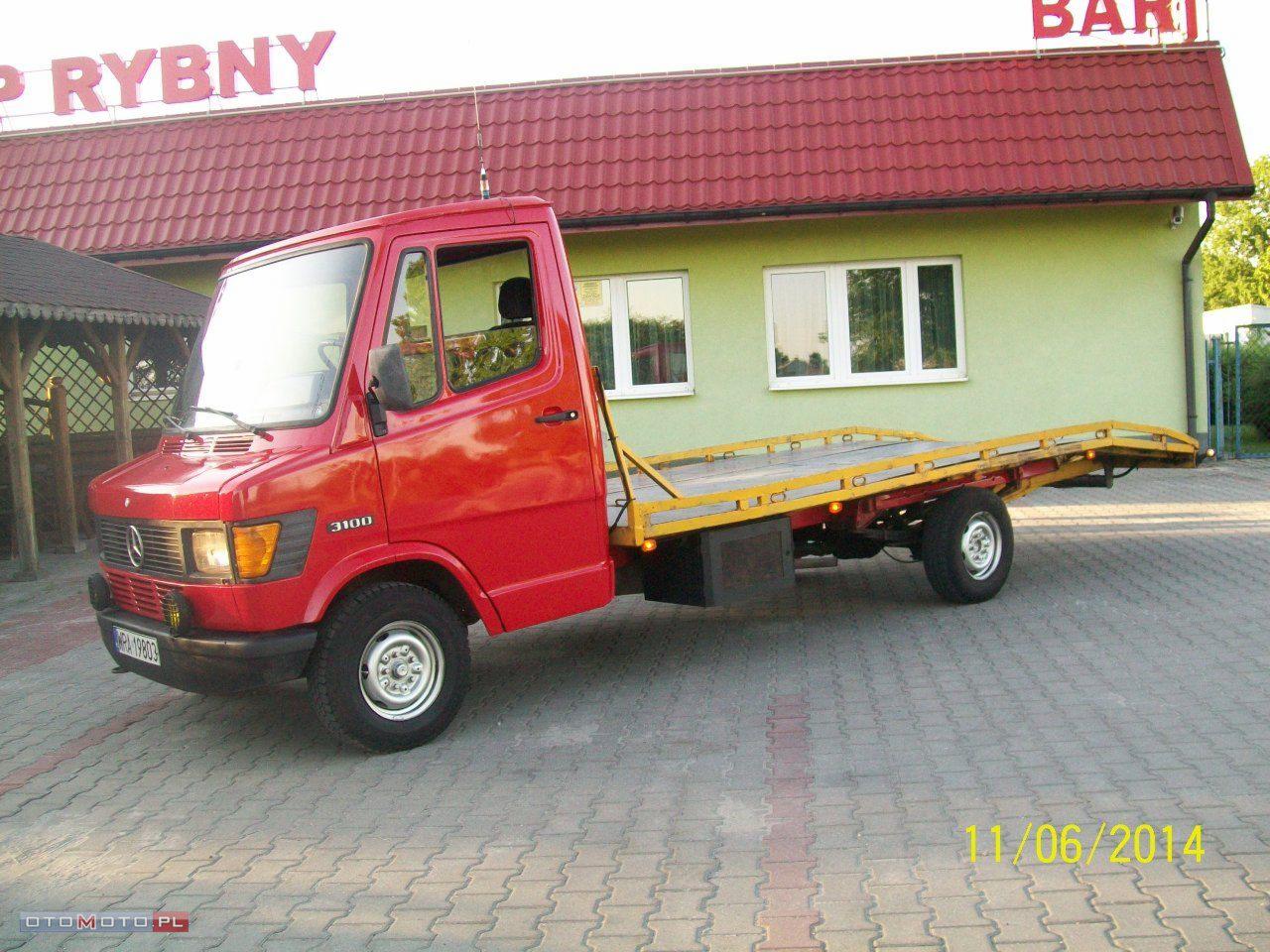 Czerwona laweta Mercedes 310D na tle budynku z czerwonym dachem. Widoczne detale: żółta platforma, opony z białymi felgami, data zdjęcia 11/06/2014.