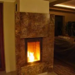 Kamienny kominek z paleniskiem, płomienie widoczne przez szybę, ciepłe światło w pomieszczeniu. Dekoracyjny element wykończenia wnętrza.