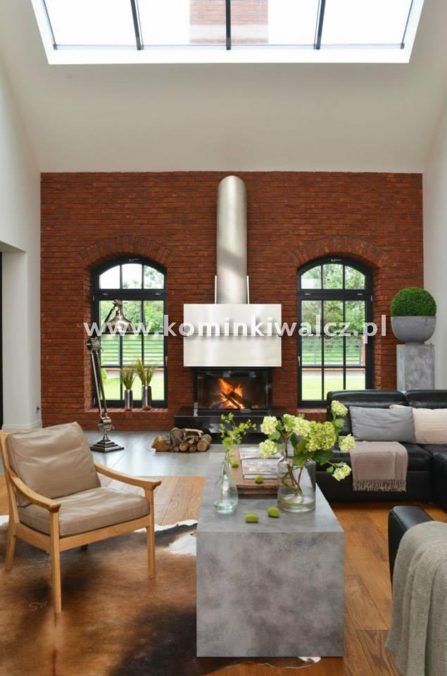 Salon z ceglaną ścianą, kominkiem z metalowym wykończeniem i dwoma oknami łukowymi. Widoczne drewniane podłogi i nowoczesne meble. W tle widać palący się ogień w kominku.