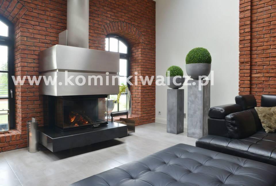 Nowoczesny kominek z metalową obudową i ceglaną ścianą w tle. Wnętrze dopełnia skórzana sofa i dekoracyjne rośliny w donicach na betonowych postumentach.