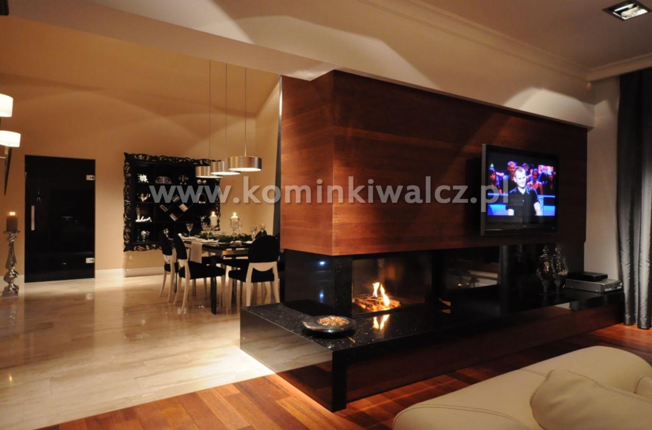 Nowoczesny salon z kominkiem obudowanym drewnem i czarnym kamieniem. Płomienie w palenisku, nad nim telewizor. W tle stół jadalny i dekoracyjna szafka.