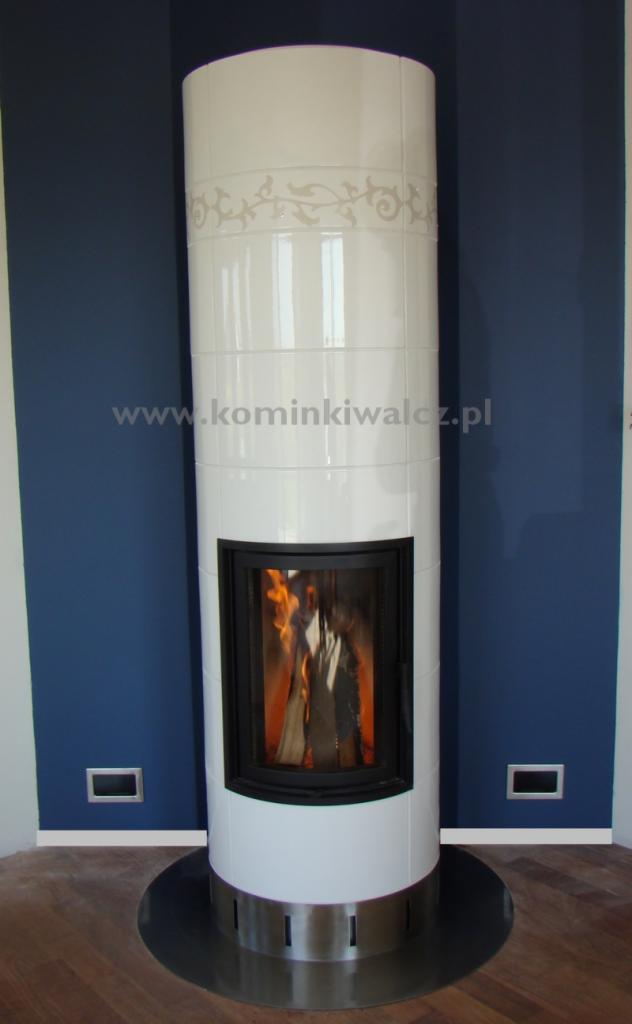 Biały, ceramiczny kominek wolnostojący z paleniskiem, zdobiony subtelnym wzorem, na tle granatowej ściany z minimalistycznymi gniazdkami.