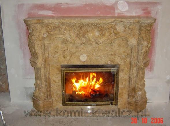 Ozdobny kominek z beżowego kamienia z płomieniami widocznymi przez szybę. Widoczne detale rzeźbienia na obudowie kominka. Częściowo wykończona ściana w tle.