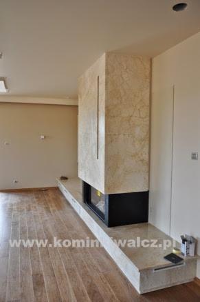 Nowoczesny kominek z obudową z beżowego marmuru, wbudowany w długi, niski blat. Minimalistyczny design w jasnym wnętrzu z drewnianą podłogą.