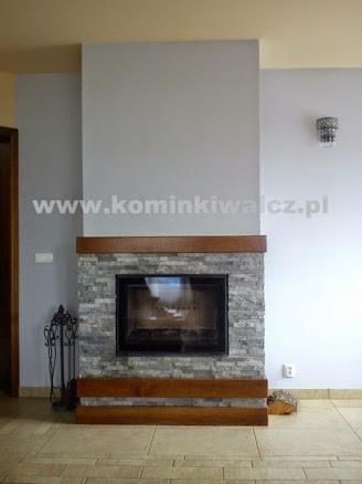 Murowany kominek z drewnianymi elementami i kamienną obudową. Widoczne akcesoria kominkowe po lewej stronie i dekoracyjny kinkiet na ścianie po prawej.